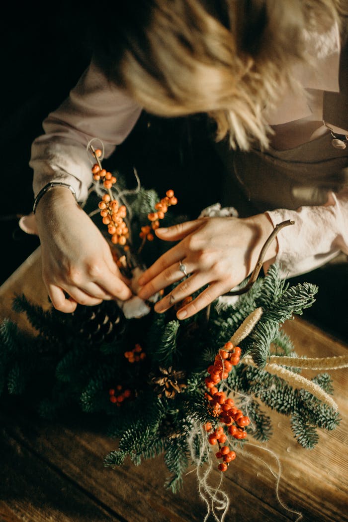 gallery-6 Hands arranging a festive Christmas wreath with pine and red berries on a wooden table.