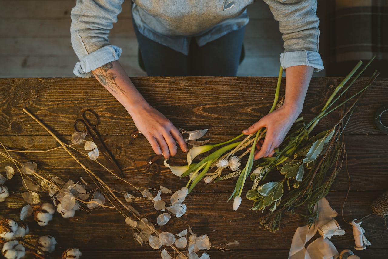 gallery-5 Tattooed hands arranging flowers on a rustic wooden table indoors, perfect for creative or floral industry scenes.
