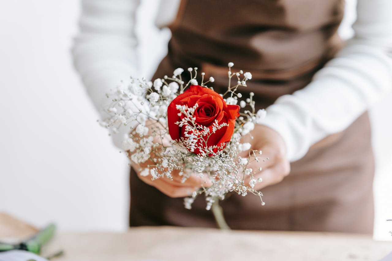 Crop unrecognizable woman florist in apron arranging bouquet with bright red rose in light room